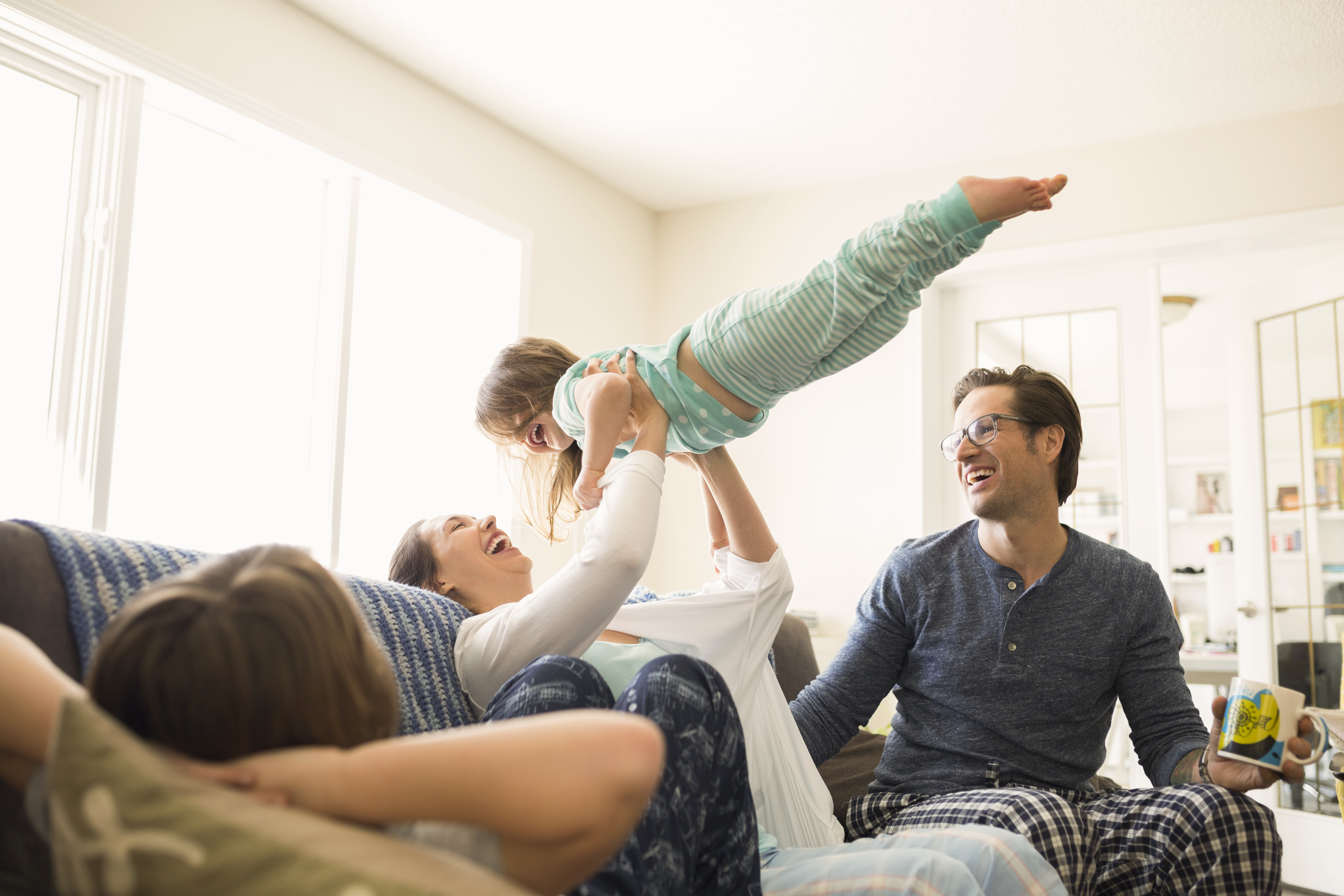 Vuxna och barn sitter i soffan iklädda pyjamas, kvinna lyfter ett barn upp i luften