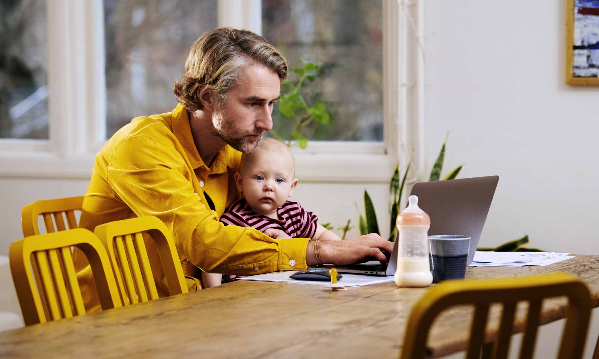 Pappa sitter hemma vid köksbordet med en bebis i knät och skriver på en laptop